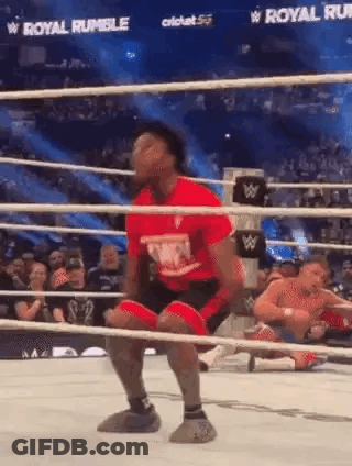 A Man In A Red Shirt Is Kneeling In A Wrestling Ring While Another Man Is Laying On The Ground . GIF