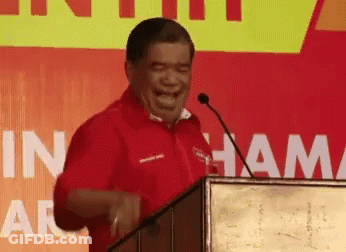A Man In A Red Shirt Is Laughing While Standing At A Podium With The Word Ham On The Wall Behind Him GIF