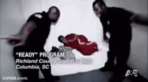 A Man In A Red Shirt Is Laying On The Floor While Two Police Officers Stand Around Him . GIF