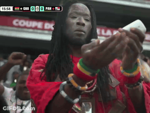 A Man In A Red Shirt Is Looking At A Cell Phone In Front Of A Scoreboard That Says Gha 0 Pan GIF