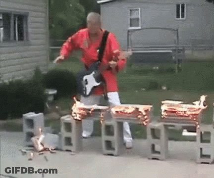 A Man In A Red Shirt Is Playing A Guitar In Front Of Some Burning Drums GIF