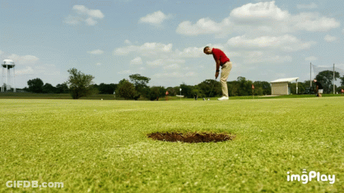 A Man In A Red Shirt Is Putting A Ball Into A Hole On A Golf Course GIF