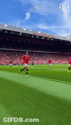 A Man In A Red Shirt Is Running On A Soccer Field In Front Of A Manchester United Stadium GIF