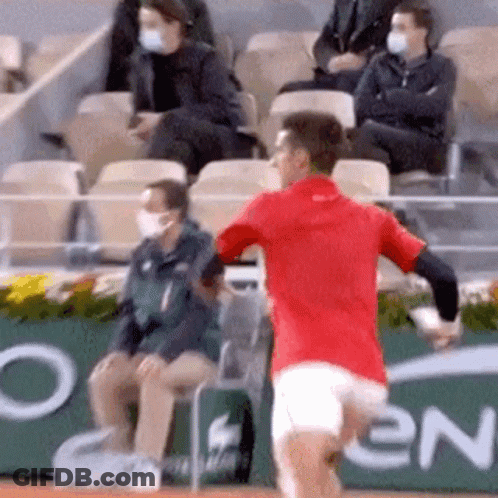 A Man In A Red Shirt Is Running On A Tennis Court In Front Of A Crowd Wearing Masks . GIF