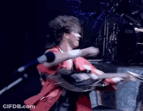 A Man In A Red Shirt Is Singing Into A Microphone In Front Of A Tama Drum Set GIF