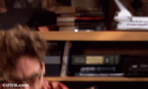A Man In A Red Shirt Is Sitting In Front Of A Bookshelf . GIF