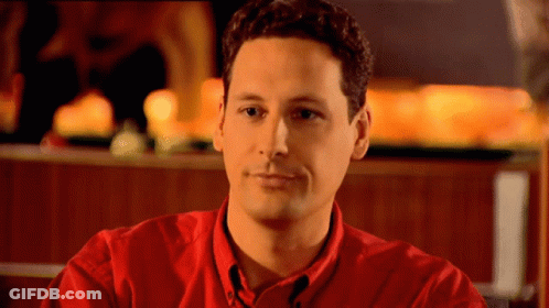 A Man In A Red Shirt Is Sitting In Front Of A Fireplace GIF