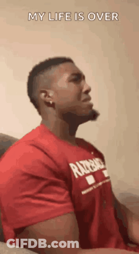 A Man In A Red Shirt Is Sitting On A Couch And Making A Face . GIF