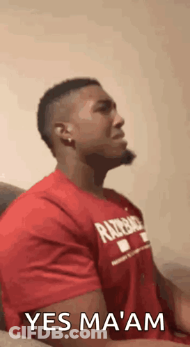 A Man In A Red Shirt Is Sitting On A Couch With His Eyes Closed And A Beard . GIF