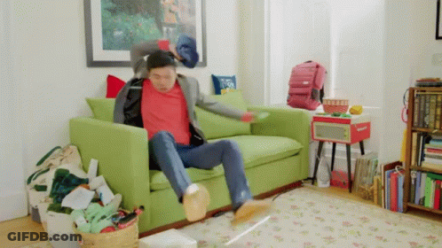 A Man In A Red Shirt Is Sitting On A Green Couch In A Messy Living Room . GIF