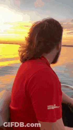 A Man In A Red Shirt Is Sitting On The Back Of A Boat Looking At The Sunset . GIF