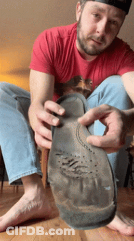 A Man In A Red Shirt Is Sitting On The Floor Holding A Shoe Sole GIF