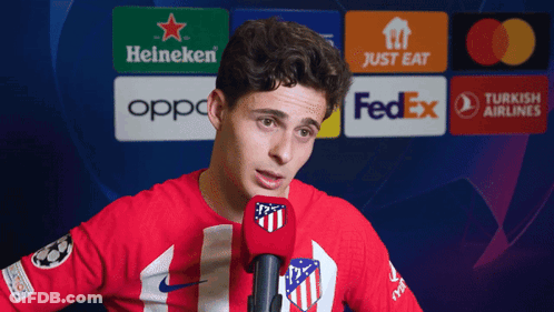 A Man In A Red Shirt Is Speaking Into A Microphone In Front Of A Heineken Logo GIF