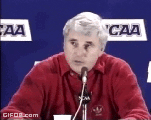 A Man In A Red Shirt Is Speaking Into A Microphone With Ncaa Logos In The Background GIF