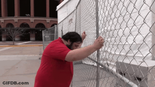 A Man In A Red Shirt Is Standing Behind A Chain Link Fence GIF