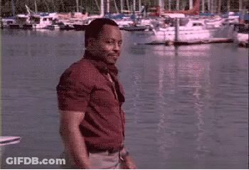 A Man In A Red Shirt Is Standing In Front Of A Body Of Water With Boats In It GIF