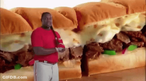 A Man In A Red Shirt Is Standing In Front Of A Very Large Sandwich . GIF