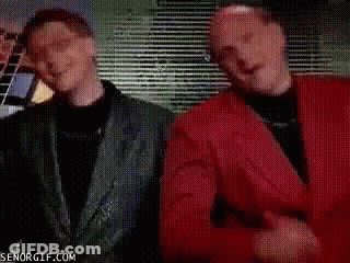 A Man In A Red Shirt Is Standing Next To Another Man In A Black Suit . GIF