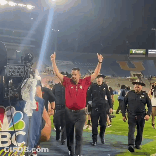 A Man In A Red Shirt Is Walking On A Field With A Nbc Sign In The Background GIF