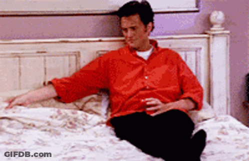 A Man In A Red Shirt Sits On A Bed GIF