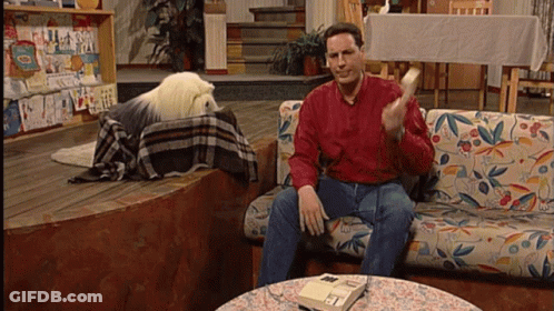 A Man In A Red Shirt Sits On A Couch With A Dog Behind Him GIF