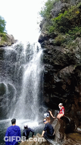 A Man In A Red Shirt Sits On A Rock Near A Waterfall GIF