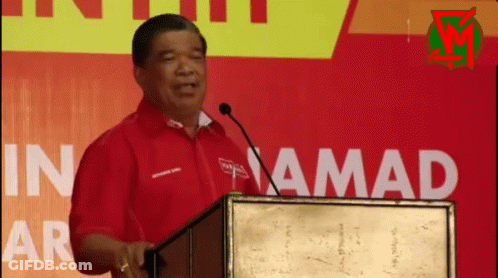 A Man In A Red Shirt Stands At A Podium In Front Of A Sign That Says In Amada GIF