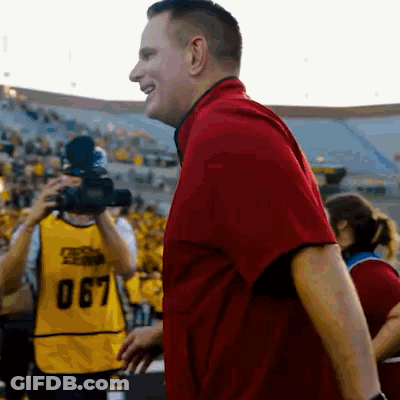 A Man In A Red Shirt Stands In Front Of A Camera Wearing A Yellow Vest With The Number 067 On It GIF