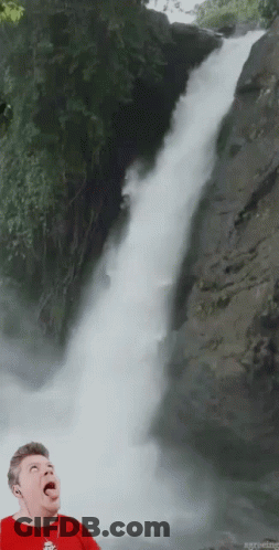 A Man In A Red Shirt Stands In Front Of A Waterfall With His Tongue Out GIF