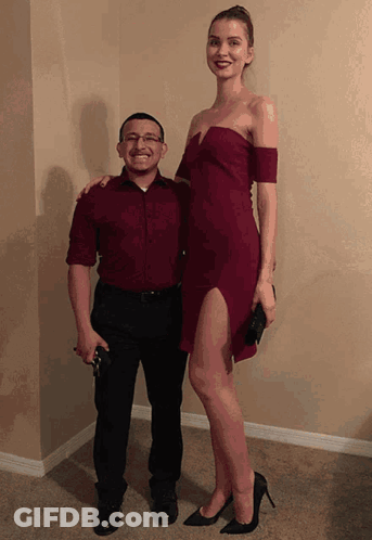 A Man In A Red Shirt Stands Next To A Very Tall Woman In A Red Dress GIF