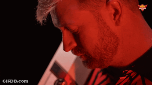 A Man In A Red Shirt With A Crown On His Ear Looks At The Camera GIF
