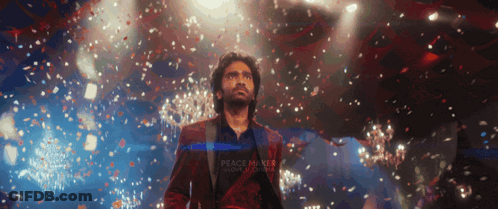 A Man In A Red Suit Stands On A Stage With Confetti Falling Around Him And A Sign That Says Peace Makes Love Stronger GIF