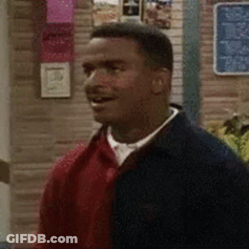 A Man In A Red Sweater And Black Jacket Is Standing In Front Of A Brick Wall . GIF