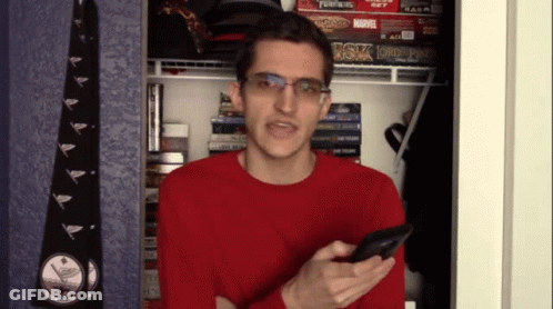A Man In A Red Sweater Is Holding A Cell Phone In Front Of A Shelf With A Board Game That Says Transformers GIF
