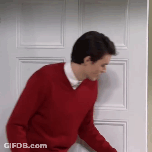 A Man In A Red Sweater Is Standing In Front Of A Door . GIF