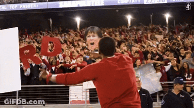 A Man In A Red Sweatshirt Stands In Front Of A Crowd Holding A Sign That Says " Cal Top Passer " GIF