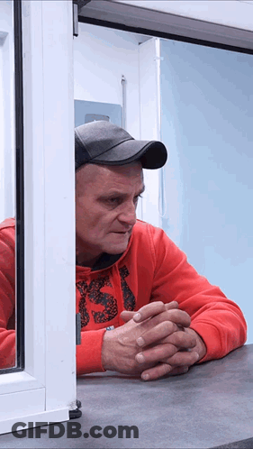 A Man In A Red Sweatshirt With The Letter S On It Looks Out Of A Window GIF
