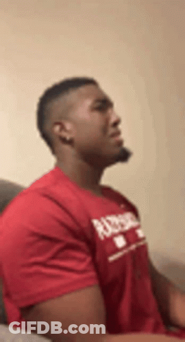 A Man In A Red T-Shirt Is Sitting On A Couch . GIF