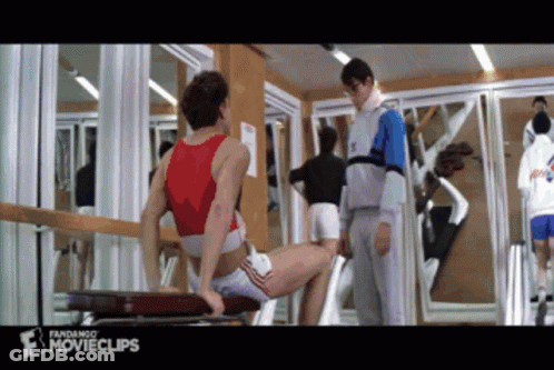 A Man In A Red Tank Top Sits On A Bench In A Gym While A Man In A Grey Jacket Stands Behind Him GIF