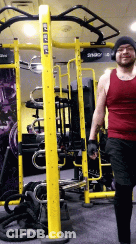 A Man In A Red Tank Top Walks In Front Of A Gym Machine That Says Synergy 360 GIF