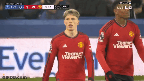 A Man In A Red Team Viewer Jersey Stands Next To Another Man On A Soccer Field GIF