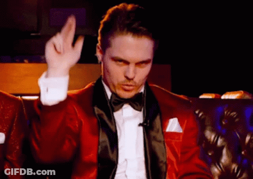 A Man In A Red Tuxedo And Bow Tie Is Sitting On A Couch Making A Gesture With His Hand . GIF