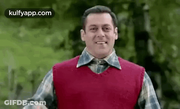 A Man In A Red Vest And Plaid Shirt Is Smiling And Looking At The Camera . GIF