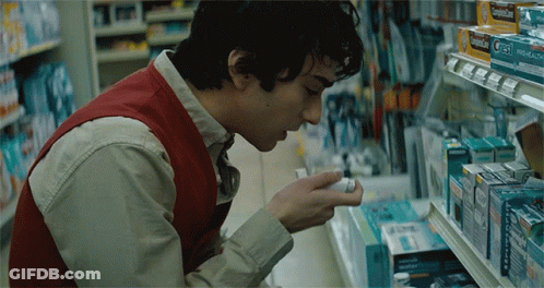 A Man In A Red Vest Is Looking At A Box Of Cold Medicine In A Store GIF