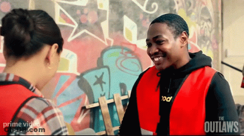 A Man In A Red Vest Is Smiling While Talking To A Woman In Front Of A Graffiti Wall . GIF