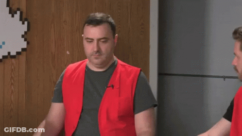 A Man In A Red Vest Is Standing Next To Another Man In A Gray Shirt . GIF