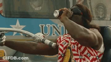 A Man In A Red White And Blue Shirt Is Drinking From A Can While Sitting In Front Of A Bus That Says Uni GIF