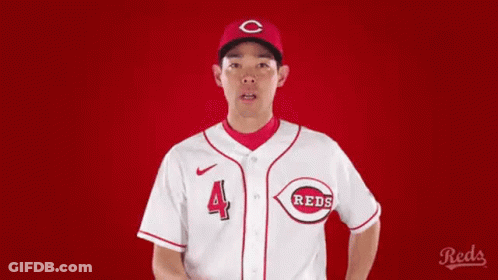 A Man In A Reds Baseball Uniform Is Pointing At Something . GIF
