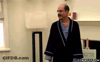 A Man In A Robe Is Standing In A Living Room With A Lamp . GIF