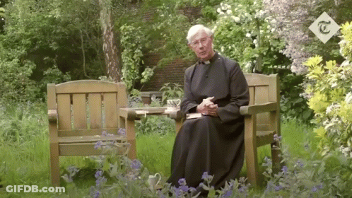 A Man In A Robe Sits In A Chair In A Garden With A T On The Corner GIF